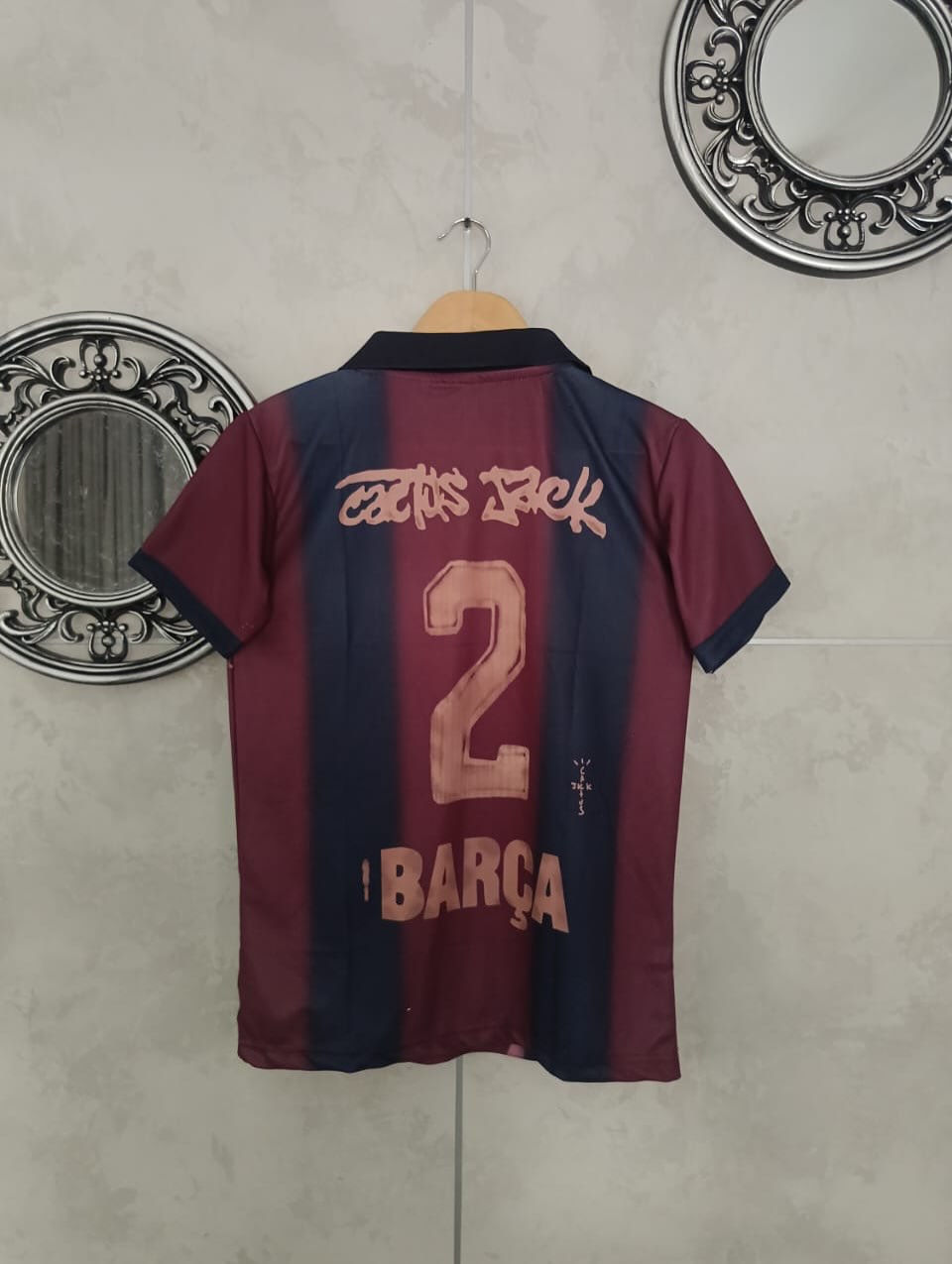 Maroon and navy sports jersey with number 2 and 'Barça' text on a hanger against a tiled wall.