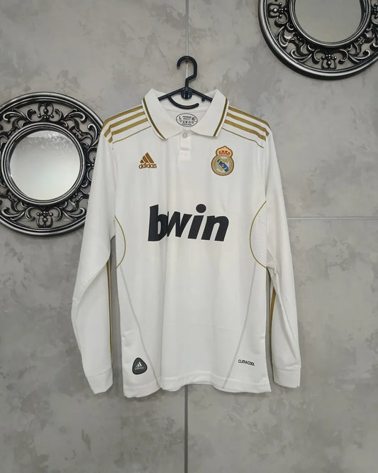White sports jersey with gold accents and brand logos on a hanger against a textured wall.